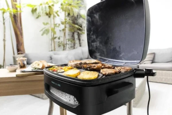 Grill elektryczny Cecotec z otwartą pokrywą, na którym smażą się warzywa i mięso, ustawiony na tarasie z meblami ogrodowymi w tle.