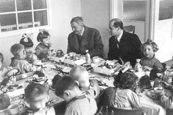 Czarno-białe zdjęcie przedstawiające Jana Wedla siedzącego przy stole z grupą dzieci podczas śniadania w 1941 roku; scena ukazuje ciepłą atmosferę spotkania z dziećmi pracowników fabryki.