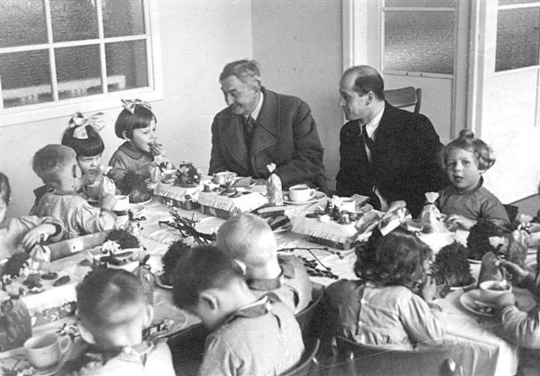 Czarno-białe zdjęcie przedstawiające Jana Wedla siedzącego przy stole z grupą dzieci podczas śniadania w 1941 roku; scena ukazuje ciepłą atmosferę spotkania z dziećmi pracowników fabryki.