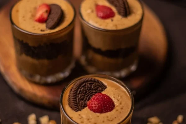 Trzy szklanki z deserem składającym się z warstw kawowego kremu (prawdopodobnie Dalgona), pokruszonych ciastek Oreo oraz udekorowane połówkami truskawek i całymi ciasteczkami Oreo. Desery stoją na drewnianej tacy, a wokół rozrzucone są okruszki. Zdjęcie przedstawia atrakcyjny deser, symbolizujący trendy i ofertę na rynku gastronomicznym w Polsce w 2025 roku.