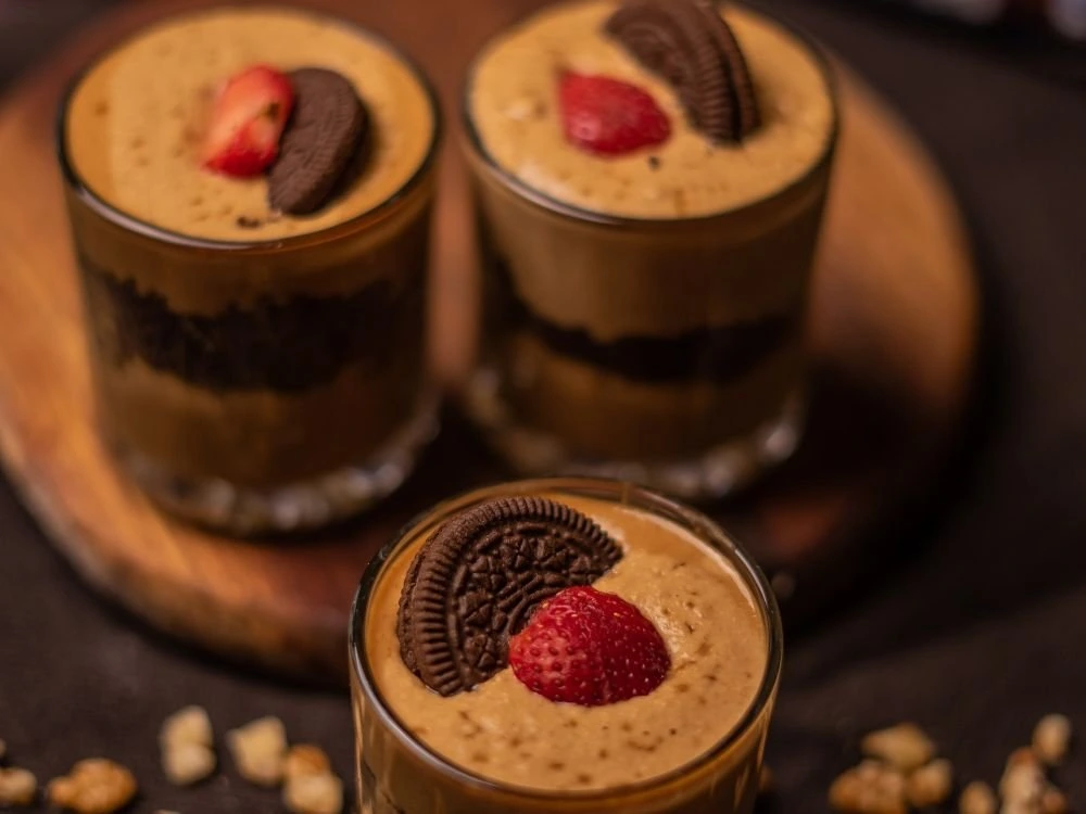Trzy szklanki z deserem składającym się z warstw kawowego kremu (prawdopodobnie Dalgona), pokruszonych ciastek Oreo oraz udekorowane połówkami truskawek i całymi ciasteczkami Oreo. Desery stoją na drewnianej tacy, a wokół rozrzucone są okruszki. Zdjęcie przedstawia atrakcyjny deser, symbolizujący trendy i ofertę na rynku gastronomicznym w Polsce w 2025 roku.