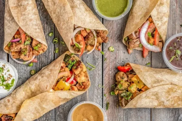 Widok z góry na pięć tortilli z różnym nadzieniem, takimi jak grillowane mięso, warzywa (papryka, cebula) i zioła, ułożonych na drewnianym stole. Obok znajdują się małe pojemniczki z różnymi sosami: zielonym (prawdopodobnie miętowym lub kolendrowym), białym (jogurtowym), brązowym (np. tamarindowym) i ciemnym (fasolowym). Obraz sugeruje różnorodność oferty charakterystyczną dla franczyzy gastronomicznej