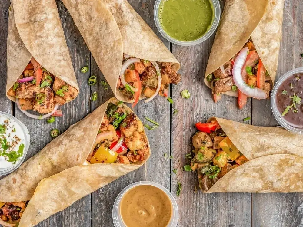 Widok z góry na pięć tortilli z różnym nadzieniem, takimi jak grillowane mięso, warzywa (papryka, cebula) i zioła, ułożonych na drewnianym stole. Obok znajdują się małe pojemniczki z różnymi sosami: zielonym (prawdopodobnie miętowym lub kolendrowym), białym (jogurtowym), brązowym (np. tamarindowym) i ciemnym (fasolowym). Obraz sugeruje różnorodność oferty charakterystyczną dla franczyzy gastronomicznej