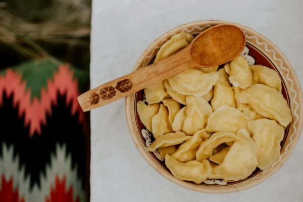 Tradycyjna miska z pierogami ustawiona na białym obrusie, z drewnianą łyżką ozdobioną ludowym wzorem.