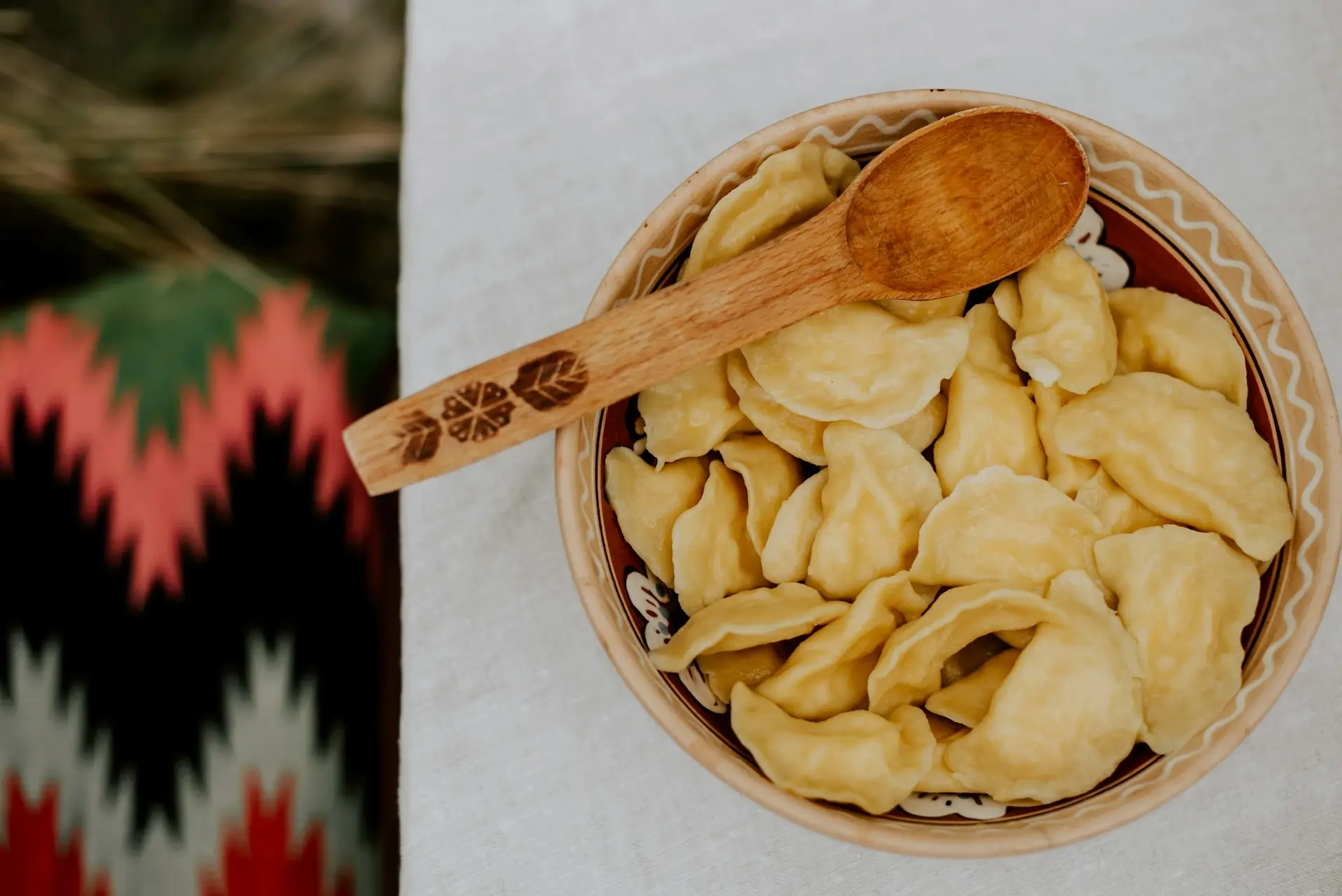 Tradycyjna miska z pierogami ustawiona na białym obrusie, z drewnianą łyżką ozdobioną ludowym wzorem.