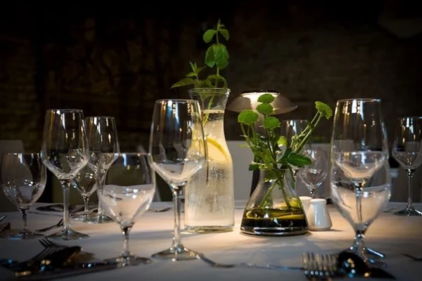 Nakryty stół w restauracji z kilkoma rzędami kieliszków do wina, sztućcami i karafką z wodą z cytryną i miętą. Obok karafki wazon z zielonymi kwiatami. Tło stanowi kamienna ściana.