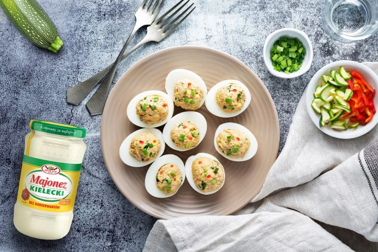 Talerz z połówkami jajek faszerowanych pastą i posypanych warzywami, obok słoik majonezu Kieleckiego, miseczki z dodatkami oraz sztućce na stole.