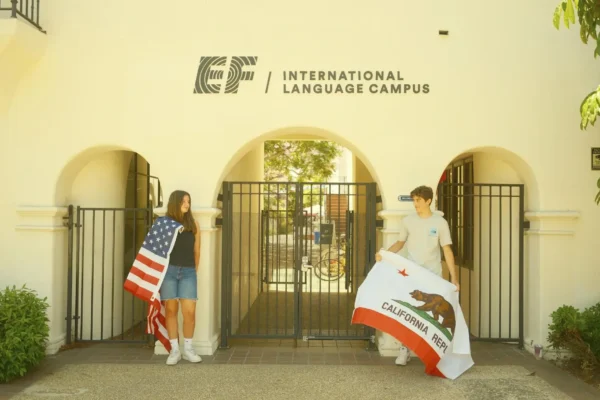Dwoje młodych ludzi stoi przed wejściem do EF International Language Campus; dziewczyna trzyma flagę USA, a chłopak flagę stanu Kalifornia.