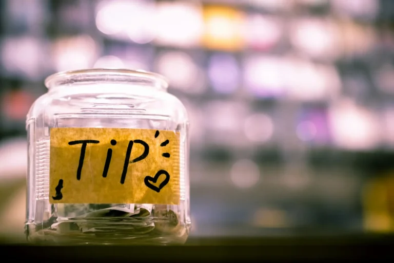 Przezroczysty szklany słoik na napiwki z naklejoną żółtą karteczką z napisem "TIP :) ♥" czarnym markerem. W słoiku znajdują się zwinięte banknoty.