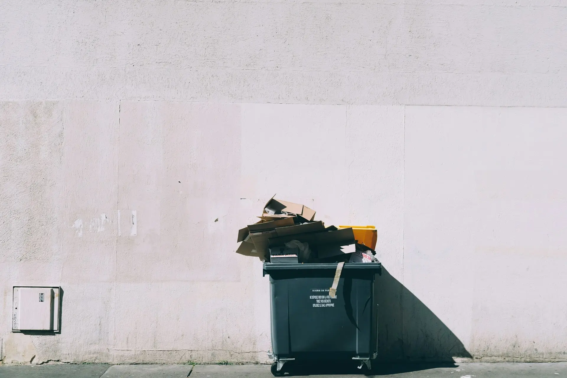 Przepełniony pojemnik na odpady stojący przy jasnej ścianie budynku, wystające kartony i inne śmieci.