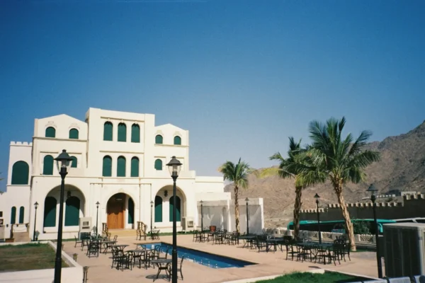 Nowoczesny, biały budynek w stylu arabskim z zielonymi oknami i łukowatymi wejściami, położony u podnóża góry. Przed budynkiem znajduje się basen, otoczony czarnymi stolikami i krzesłami oraz palmami.
