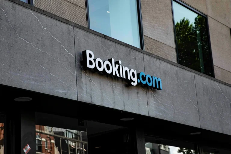 Zewnętrzny widok na budynek o szarym, marmurowym wykończeniu. Nad wejściem widnieje trójwymiarowe, podświetlane logo serwisu Booking.com.