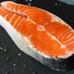 Duży, świeży, surowy stek z łososia firmy suempol leży na ciemnej kamiennej płycie. Wokół steka rozsypana jest gruboziarnista sól i kilka ziaren czarnego pieprzu.